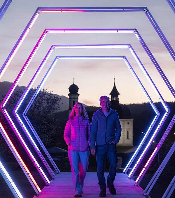 lichterpark-seefeld-2024-mann-und-frau-gehen-durch-lichtertunnel-mit-seekirchl-im-hintergrund-1