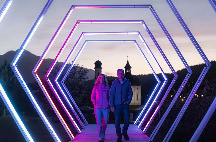 lichterpark-seefeld-2024-mann-und-frau-gehen-durch-lichtertunnel-mit-seekirchl-im-hintergrund